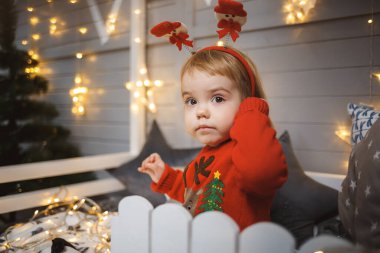 Kırmızı, sıcak kazaklı küçük bir kız Noel ağacının yanındaki bir yatakta oyuncak ve hediyelerle oturuyor. Mutlu bir çocukluk. Yeni yıl tatili atmosferi