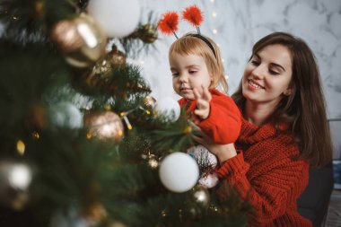 Kırmızı süveter giyen anne ve kız Noel ağacını süslüyor. Mutlu annelikler. Sıcak aile ilişkileri. Noel ve Yeni Yıl arifesi. Sevgi. Aile kavramı.