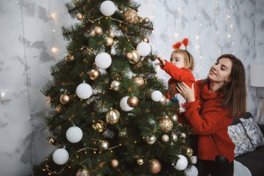 Kırmızı süveter giyen anne ve kız Noel ağacını süslüyor. Mutlu annelikler. Sıcak aile ilişkileri. Noel ve Yeni Yıl arifesi. Sevgi. Aile kavramı.