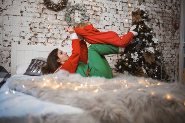 Kırmızı, sıcak kazaklı kızı olan anne yatakta zıplıyor. Mutlu annelikler. Sıcak aile ilişkileri. Noel ve Yeni Yıl arifesi. Sevgi. Aile kavramı.