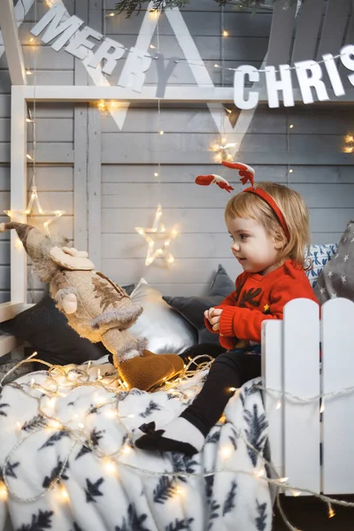 A little girl in a red warm sweater sits on a bed near a Christmas tree with toys and gifts. Happy childhood. New Year holiday atmosphere