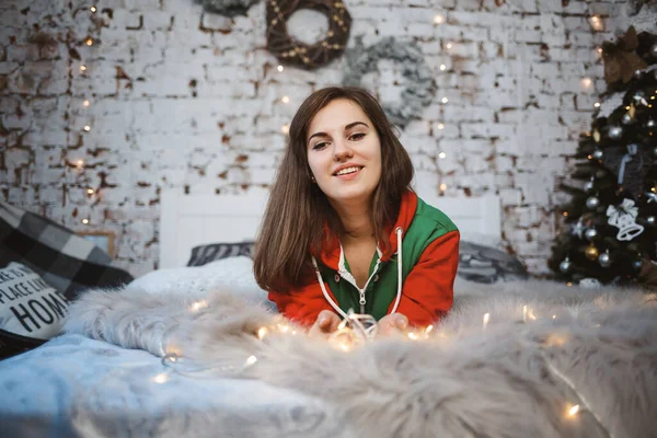 Dziewczyna Noworocznym Kombinezonie Świątecznym Humorem Leży Łóżku Wokół Świecących Girland — Zdjęcie stockowe