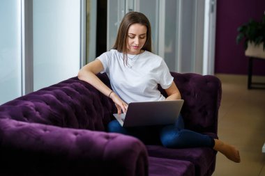 Genç bir kadın kanepede oturuyor ve işten uzak bir dizüstü bilgisayarla çalışıyor. Dizlerinin üstünde bilgisayarı olan bir kız monitöre bakıyor.