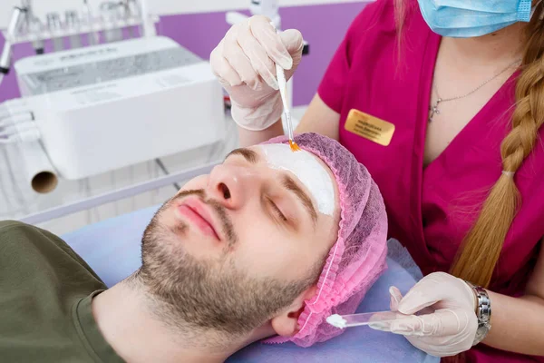 Cosmetologist is applying white moisturizing mask on man\'s face using brush in beauty clinic, face closeup. Beauty industry concept. Guy is lying on couch. Beautician woman making spa procedure.