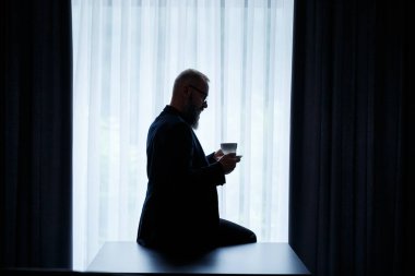 Sakallı yetişkin bir adam kahve içer ve pencereden dışarı bakar. Çalışma günü eğlence konsepti