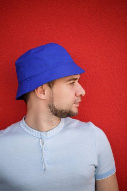 Kafasında panama şapkası olan sakallı genç yakışıklı bir adam kırmızı bir duvarın arkasında. Beyaz tişörtlü bir adamın portresi.