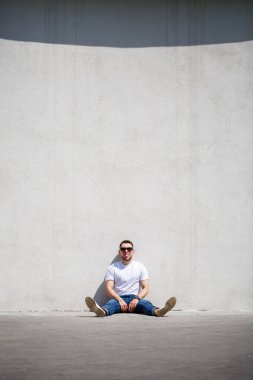 Tişörtlü sakallı genç bir adam gri bir duvarın yanında oturuyor. Güneş gözlüklü adam.