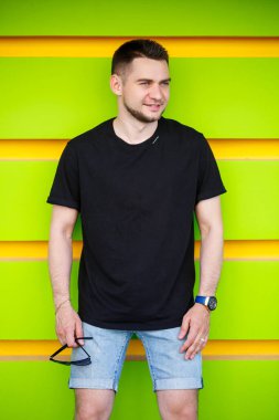 Siyah tişörtlü ve şortlu parlak yeşil arka planda genç bir adam.