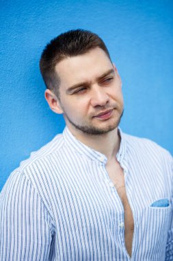 Gömlekli, sakallı yakışıklı bir adam mavi arka planda duruyor..