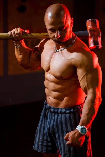 Strong athletic man posing with a sports hammer on the background of the gym. A strong bodybuilder with perfect abs, shoulders, biceps, triceps and chest.