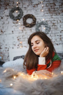 Yeni yıl tulumu giymiş neşeli bir kız parlak çelenklerin etrafında bir yatakta yatıyor.