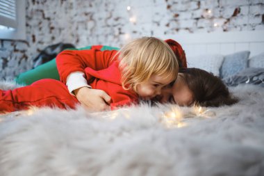 Anne ve kızı, kırmızı, sıcak süveter giymiş yatağın üstünde. Mutlu annelikler. Sıcak aile ilişkileri. Noel ve Yeni Yıl arifesi. Sevgi. Aile kavramı.