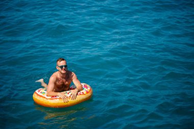 Genç adam güneşli bir günde denizde şişme bir yüzükte dinleniyor ve yüzüyor.