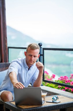 Başarılı bir erkek işadamı, dağ manzaralı bir dizüstü bilgisayarın arkasında tatilde çalışıyor. Çevrimiçi yönetici iş akışı. Balkondan güzel bir manzarayla dışarıda çalışmak.