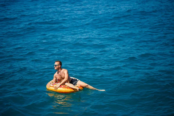 一个年轻人在大海中的一个充气气圈上漂浮着蓝色的水 在阳光明媚的日子里过节 — 图库照片