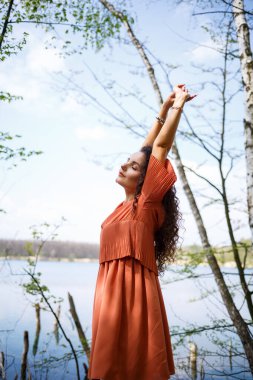 Yüzünde gülümseme, mutlu duygular olan genç bir kadın. Uzun dalgalı kıvırcık saçlı bir kız yazın doğada, gölde bir ormanda, bir yolun kenarında ve çalıların arasında duruyordu.