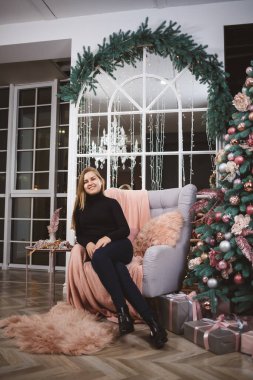 Örgü kazaklı güzel bir kadın Noel ağacıyla süslenmiş lüks bir odanın arka planında bir koltuğa oturur. İyi yeni yıl ruhu.