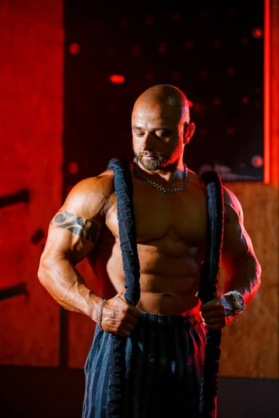 Sporty handsome strong man posing with sports rope on the background of the gym. A strong bodybuilder with perfect abs, shoulders, biceps, triceps and chest.