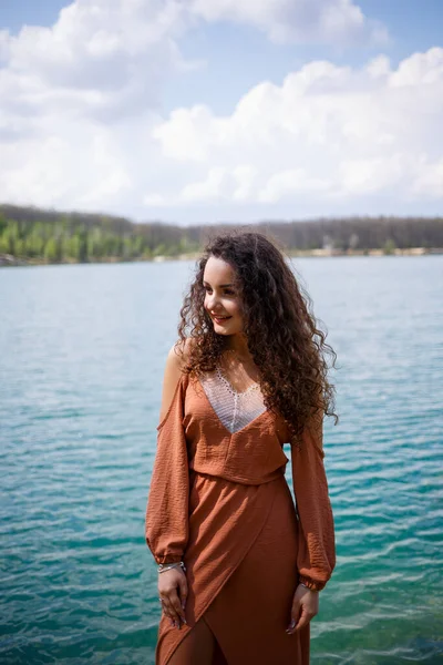 青い湖の真ん中にある木製の石積みの上に若い女性が立っている 女の子幸せな笑顔と太陽の輝き 夏の日 彼女は巻き毛とヨーロッパの外観を持っています — ストック写真