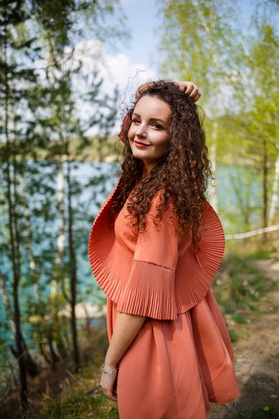 Wood masonry. A girl with long wavy curly hair in an orange guipure dress and shoes on nature, in a forest by the lake, stood near trees and bushes. Young woman smiles and enjoys life