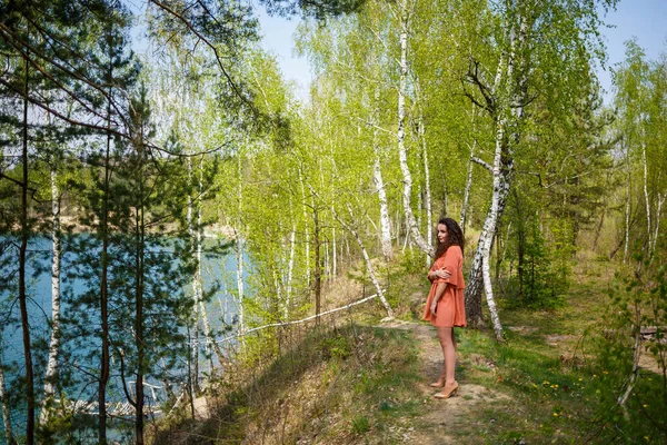 Wood masonry. A girl with long wavy curly hair in an orange guipure dress and shoes on nature, in a forest by the lake, stood near trees and bushes. Young woman smiles and enjoys life
