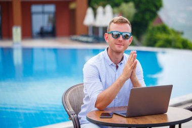 Güneş gözlüklü genç erkek işadamı havuzun yanındaki masada oturan bir dizüstü bilgisayarda çalışıyor. Uzaktan kumanda. Serbest Çalışan