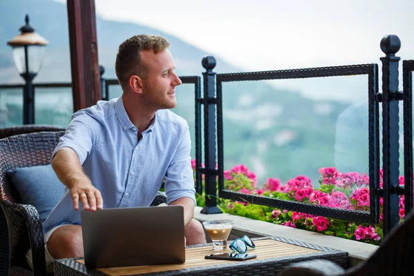 Successful male businessman working on vacation behind a laptop with a mountain view. Online manager workflow. Work outdoors with a beautiful view from the balcony