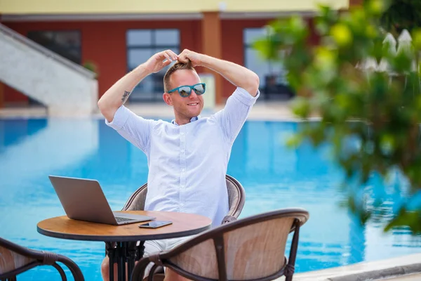 Güneş gözlüklü başarılı yakışıklı iş adamı havuz kenarında oturan bir dizüstü bilgisayarda çalışıyor. Uzaktan kumanda. Serbest Çalışan