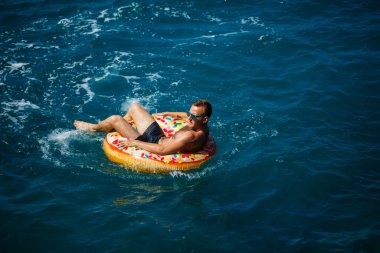 Genç bir adam güneşli bir günde şişme bir yüzükle açık denizde yüzer. Yaz tatili, tatildeki turist.