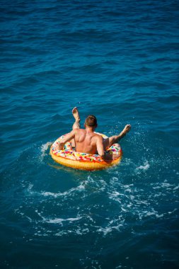 Genç bir adam mavi suyla denizde şişirilebilir bir halka üzerinde yüzüyor. Güneşli bir günde güzel bir tatil. Tatil konsepti, üst görünüm.