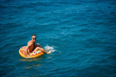 Genç bir adam mavi suyla denizde şişirilebilir bir halka üzerinde yüzüyor. Güneşli bir günde güzel bir tatil. Tatil konsepti, üst görünüm.