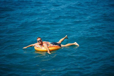 Genç bir adam mavi suyla denizde şişirilebilir bir halka üzerinde yüzüyor. Güneşli bir günde güzel bir tatil..