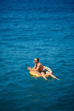Genç bir adam mavi suyla denizde şişirilebilir bir halka üzerinde yüzüyor. Güneşli bir günde güzel bir tatil..