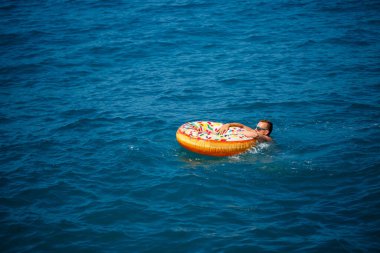 Genç bir adam mavi suyla denizde şişirilebilir bir halka üzerinde yüzüyor. Güneşli bir günde güzel bir tatil..