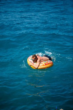 Genç adam güneşli bir günde denizde şişme bir yüzükte dinleniyor ve yüzüyor.