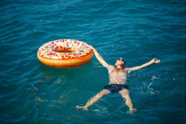 Genç adam güneşli bir günde denizde şişme bir yüzükte dinleniyor ve yüzüyor.