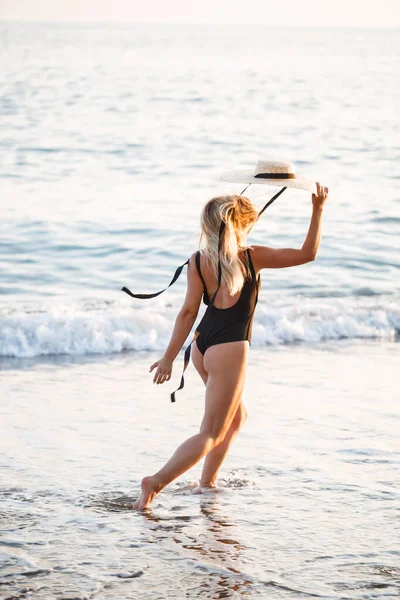日没の日差しの中で海で黒い水着と砂浜の帽子の美しい女の子 — ストック写真