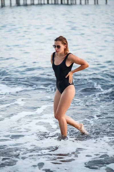 海によってビーチにポーズブロンドの髪と黒の水着で美しい女性の肖像画 ビーチを歩いている若い女性 — ストック写真