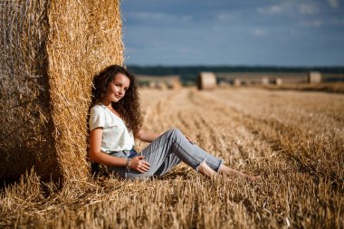 Tarlada saman demetinin yanında güzel bir genç kadın. Köyde tatiller, güneşli bir günde çim biçme tarlasında eğlenen bir kız.