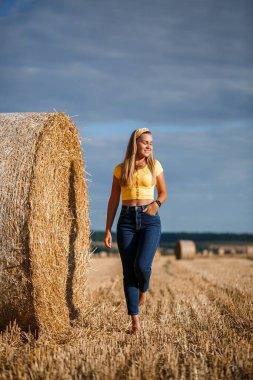 Genç, güzel bir sarışın, biçilmiş buğday tarlasında büyük bir saman demetinin yanında duruyor ve doğanın tadını çıkarıyor. Köyün doğası.