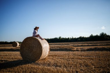 Şapkalı ve yazlık elbiseli güzel bir kadın samanlıkta samanlıkta oturuyor. Kırsal doğa, buğday tarlası