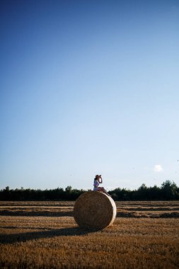 Şapkalı ve yazlık elbiseli güzel bir kadın samanlıkta samanlıkta oturuyor. Kırsal doğa, buğday tarlası