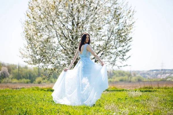 花の木に対して彼女の髪に飾られた明るい夏の青い長いドレスの女の子の肖像画 — ストック写真