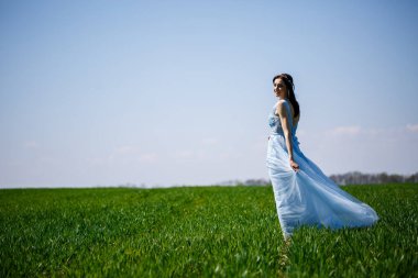 Yeşil tarlada, mavi, uzun elbiseli bir kadın. Yüzünde bir gülümseme olan güzel bir kızın moda portresi.