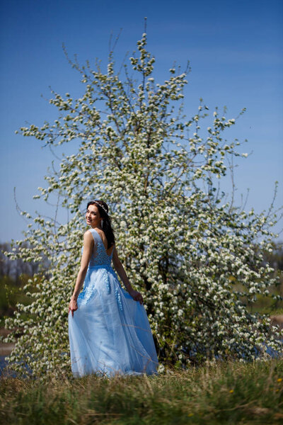 Portrait of a tender girl in a blue long dress under a blossoming tree with a smile on her face on a sunny warm summer day