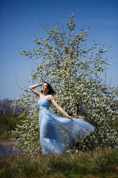 暖かい夏の日に彼女の顔に笑顔と花の木の下の青い長いドレスで柔らかい女の子の肖像画 — ストック写真
