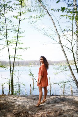 Yüzünde gülümseme, mutlu duygular olan genç bir kadın. Uzun dalgalı kıvırcık saçlı bir kız yazın doğada, gölde bir ormanda, bir yolun kenarında ve çalıların arasında duruyordu.