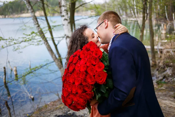 年轻人的浪漫聚会 一个年轻的女人同意嫁给她的男人 一个穿着西服 捧着一束红玫瑰的男人送给女孩一束红玫瑰 女孩们在森林里接吻 — 图库照片