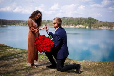 Açık havada bir kadının eline gümüş bir alyans takan bir adam resmi. Deniz ya da nehir arkaplanı