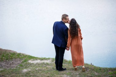 Mavi gölün arka planında el ele tutuşan genç bir çift. Uzun elbiseli bir kız ve takım elbiseli bir erkek. Doğa, manzara. Aşk ve aşk, mutlu çift